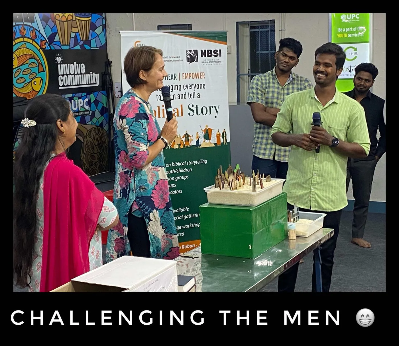A woman speaks to a group of men and women in front of a display, captioned Challenging the men.