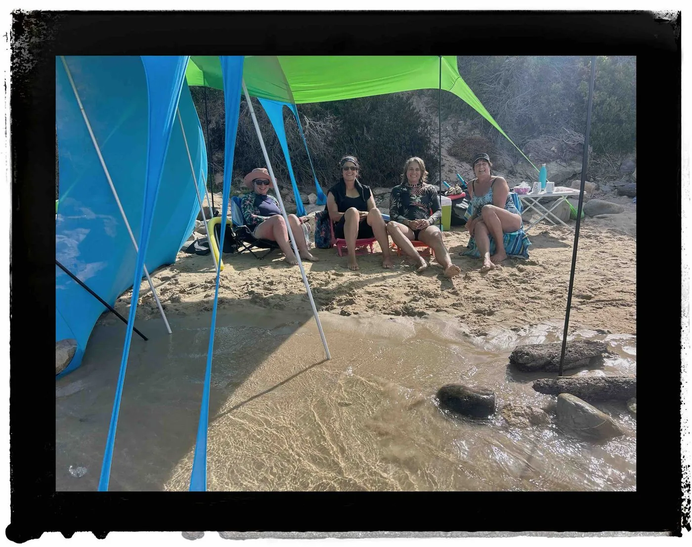 A group of people sitting under a blue tarp shelter on a beach.
